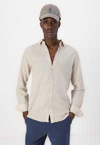 Camisa de lino beige con frente abotonado y puños enrollados, combinada con pantalones azul marino. La gorra es de un gris claro con un diseño bordado.