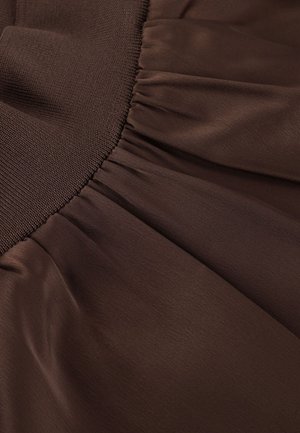 Brauner, leichter Stoff mit glatter Textur. Verfügt über gesammelte Falten in der Nähe eines gerippten Bunds, die fließende Formen und Tiefe erzeugen.