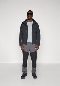 Black waterproof jacket with multiple pockets, paired with grey and black color-blocked athletic pants and black rugged shoes.