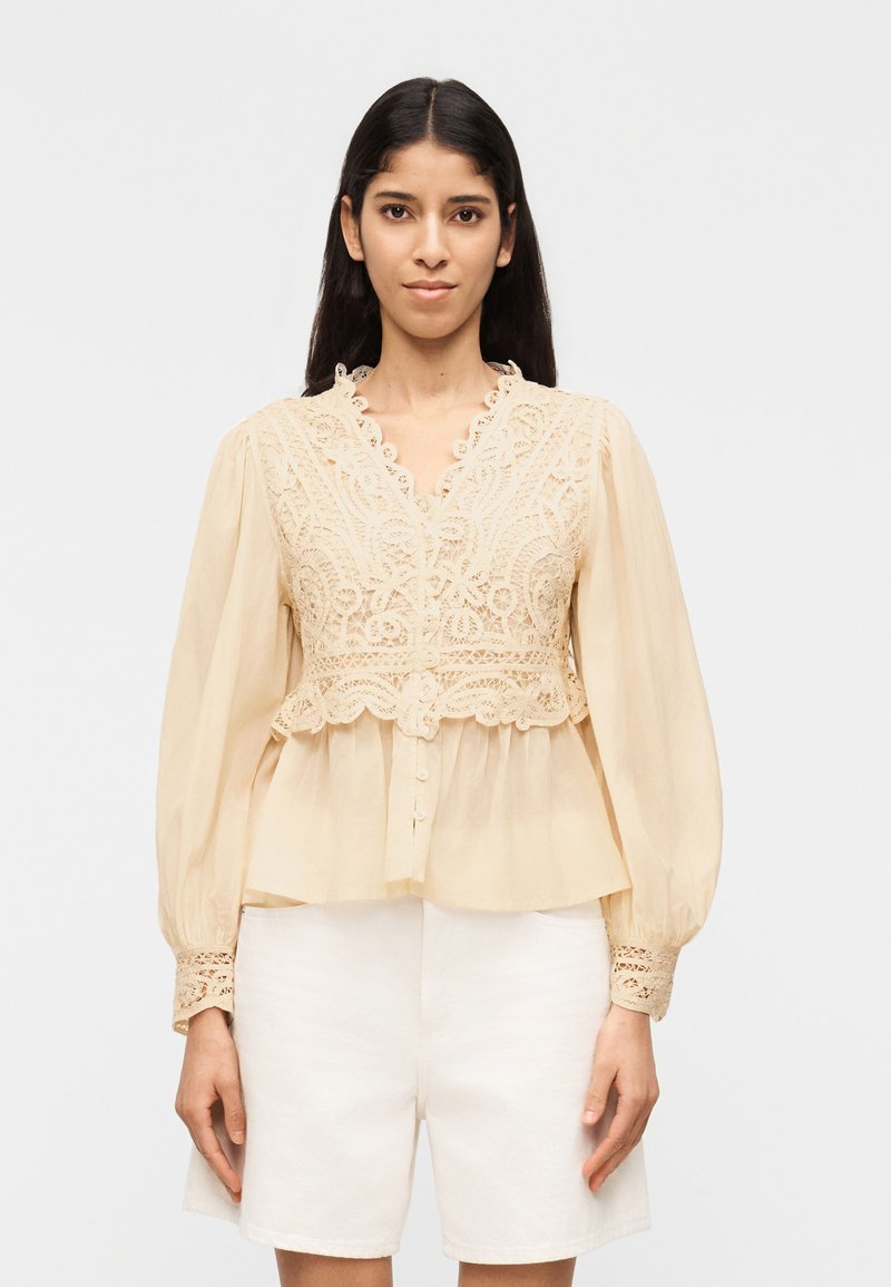 Γυναίκα που φοράει μπεζ μπλούζα με δαντελωτές λεπτομέρειες και μακριά μανίκια με κουμπιά, και λευκά σορτς ψηλόμεσα, στέκεται μπροστά σε απλό φόντο.