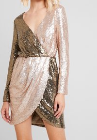 Robe enveloppante en sequins avec un décolleté en V, des manches longues et un ourlet asymétrique. Les couleurs varient du doré au rose clair. Doublure intérieure douce.