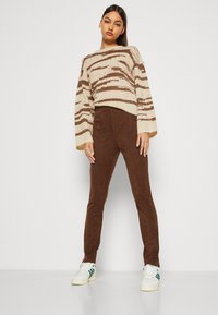 Strikkegenser med kremfargede og brune striper, kombinert med brune bukser i nubuck-liknende materiale og hvite joggesko med grønne detaljer.