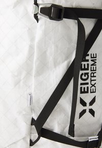 Rucsac din țesătură albă cu un model în zig-zag, curele și accesorii negre. Include logo-uri ale brandului și detalii ale buzunarului exterior.