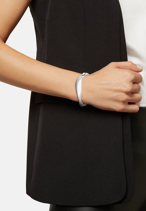 Siliconen armband in lichtgrijs, dun en soepel ontwerp, vastgemaakt met een minimalistische sluiting, gedragen om een pols tegen een donkere kledingstuk.