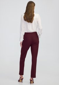 Vínové kalhoty na míru s hladkou texturou, elastickým pasem a úzkým střihem, doplněné bílou blůzou s dlouhým rukávem a podpatky.