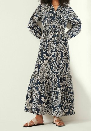 Navy blue paisley-patterned maxi dress with long sleeves, button front, and tiered skirt. Worn with light sandals.