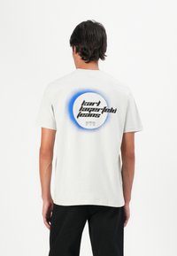 Mand iført en hvid T-shirt med et blåt cirkulært grafisk design og sort tekst "Karl Lagerfeld Jeans" på ryggen, parret med sorte bukser.