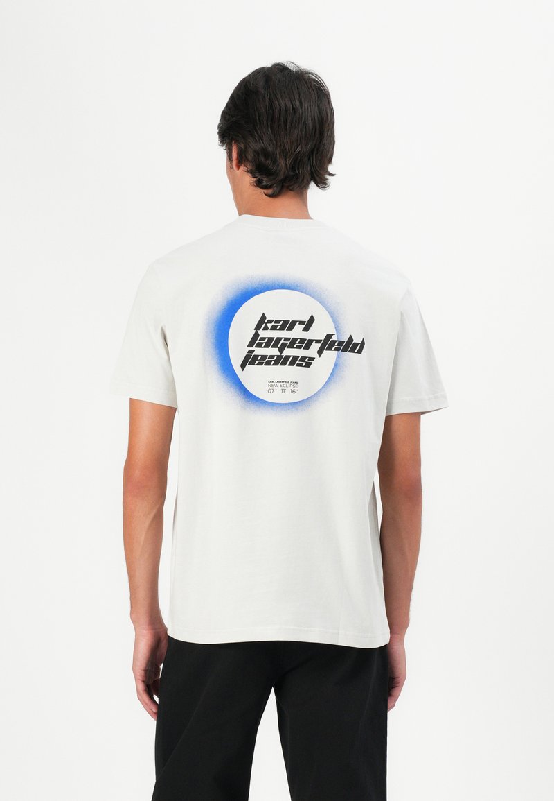 Mand iført en hvid T-shirt med et blåt cirkulært grafisk design og sort tekst "Karl Lagerfeld Jeans" på ryggen, parret med sorte bukser.