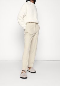 Witte geribde trui, beige op maat gemaakte broek en lichtgrijze instappers met bruine accenten. Minimalistisch ontwerp, zachte texturen en een relaxte pasvorm.