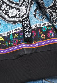 Versace Jeans Couture-produkt med ett blommönster på svart tyg, kontrasterande denim och en framträdande svart etikett med vit text.