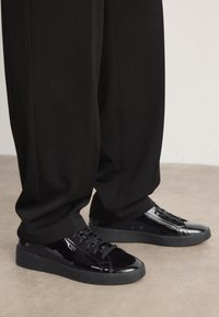 Zapatillas de deporte de charol negro con acabado suave, punta redonda y suelas de goma planas. Sin logos visibles; los cordones son negros y texturizados.