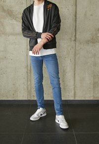 Ung person i svart Nike-jacka, vit t-shirt, ljusblå jeans och vita Nike-sneakers, stående mot en betongvägg.