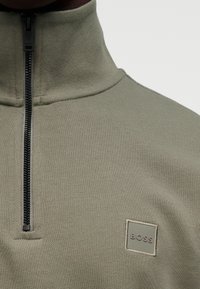 Πουλόβερ φλις σε ελιά πράσινο με ψηλό γιακά, εμπρόσθιο φερμουάρ και μικρό τετράγωνο ένθετο λογότυπο με την επιγραφή "BOSS" στο αριστερό στήθος.