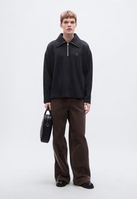 Sweat-shirt noir à zip avec un col montant, associé à un pantalon marron ample. Le modèle tient un sac à main noir. Texture lisse, design minimaliste.