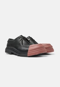 Zapatos de cuero negro con una puntera de goma rosa sólido, suela plana negra y un sutil detalle de cordones. Textura suave y forma redondeada.