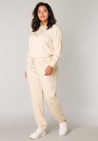 Beige loungewear set met een hoodie met trekkoorden en bijpassende joggers. Glad materiaal met een relaxte pasvorm en zijzakken. Witte sneakers.