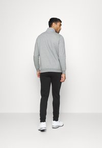 Felpa grigia con colletto alto, pantaloni neri e scarpe sportive bianche. Tessuto liscio con polsini e orlo a costine. Logo del marchio visibile sulle scarpe.