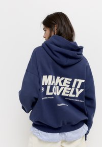 Marineblauwe hoodie met een grote witte grafische tekst op de achterkant, met de tekst "MAKE IT LOVELY." Gemaakt van zachte stof, met een losse pasvorm en zak.