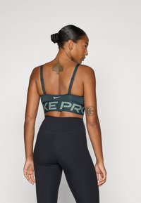 Temno zelena športna nedrčka z nastavljivimi naramnicami, ki ima širok pas s napisom "NIKE PRO". Nosi se z črnimi leginami z visokim pasom.