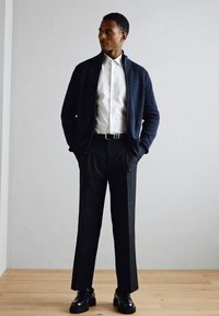 Maglione navy a coste con zip, camicia bianca, pantaloni neri e scarpe nere, posati su un pavimento in legno con uno sfondo neutro.