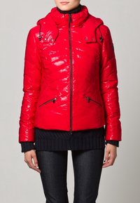 Röd puffy jacka med blank finish, dragkedja fram, justerbar huva och sidofickor. Bärs över en mörk stickad tröja och jeans.