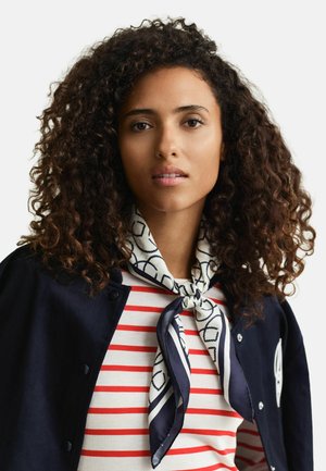 Jonge vrouw met krullend donker haar, gekleed in een rood-wit gestreept shirt, een marineblauwe jas en een zijden sjaal met patroon om haar nek geknoopt.