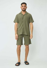 Chemise à manches courtes vert olive et shorts assortis en texture côtelée. Sandales noires à enfiler. Look décontracté et ample.