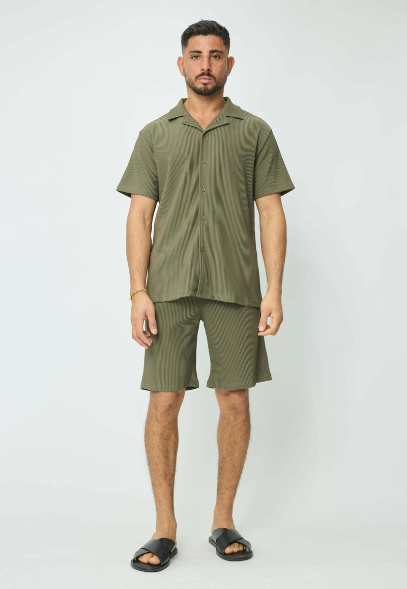 Chemise à manches courtes vert olive et shorts assortis en texture côtelée. Sandales noires à enfiler. Look décontracté et ample.