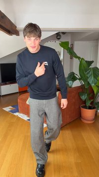 Νεαρός άνδρας με καστανά μαλλιά που φοράει μαύρο φούτερ και γκρι παντελόνι δείχνει ένα λογότυπο στο στήθος του σε ένα μοντέρνο σαλόνι.