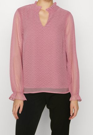 Frau trägt eine transparente, rosa, strukturierte Bluse mit langen Ärmeln und Rüschensäumen an Manschetten und Ausschnitt, kombiniert mit schwarzen Hosen.