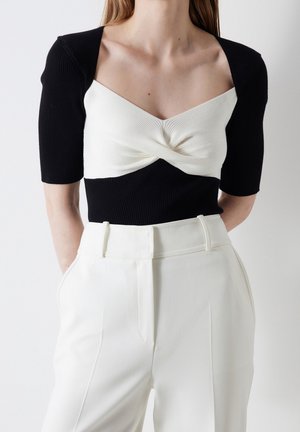 Kobieta ubrana w czarno-biały dzianinowy top z przednim skrętem, rękawami do łokcia oraz białe spodnie z wysokim stanem na jednolitym tle.