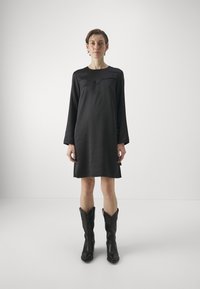 Abito nero a maniche lunghe con scollatura curva e texture liscia, abbinato a stivali da cowboy neri alti con punta affusolata.