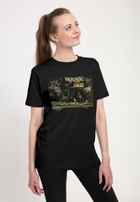 Žena s dlouhými vlasy, oblečená v černém tričku s grafikou starožitného kola s košíkem na pozadí ve stylu graffiti.