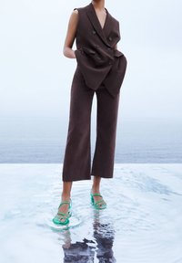 Brown double-breasted sleeveless vest with pockets, paired with brown cropped trousers and green strappy sandals, standing in shallow water.