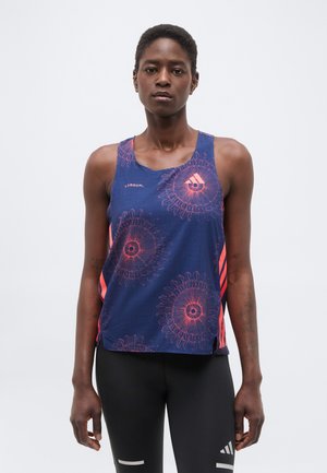 Γυναίκα που φοράει μπλε αθλητικό τοπ χωρίς μανίκια με ροζ κυκλικά σχέδια και μαύρο κολάν με λευκές ρίγες, στέκεται μπροστά σε απλό φόντο.