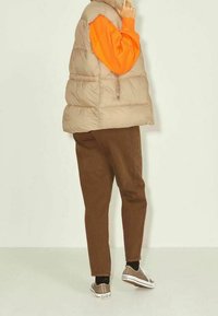 Blandad beige väst över en orange tröja, bruna byxor och beige sneakers. Slät textur, dragkedjedetalj och avslappnad passform.