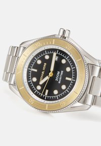 Reloj de acero inoxidable con cara negra, bisel dorado y acentos plateados. Presenta marcadores de hora redondos y una ventana de fecha a las 3 en punto.