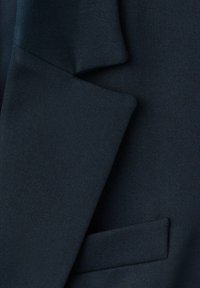 Marineblauwe op maat gemaakte jas van stof met een scherpe reverskraag, een gestructureerde weving en een enkele paspelzak, die strakke lijnen en een gestructureerd ontwerp benadrukt.