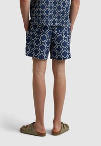 Shorts de bain bleu marine avec un motif géométrique blanc, présentant une coupe décontractée et des poches latérales. Associés à des sandales olive.