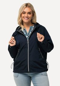 Marinblå vindjacka med huva, med dragkedja fram, två sidofickor och justerbara dragsnören vid nederkanten. Slät textur.