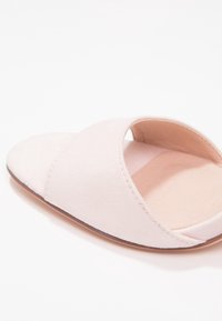 Avoin varvas liukusandaali pehmeästä vaaleanpunaisesta kankaasta, jossa on ristikkäinen malli ja sileä, beige sisäpohja littealla pohjalla.