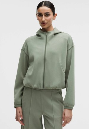 Zelená mikina na zips vyrobená z mäkkej látky, s voľným strihom, kapucňou, bočnými vreckami a rebrovanými manžetami. Hladká textúra s minimálnymi detailmi.