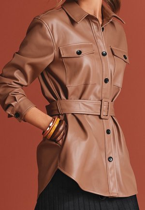 Chemise en cuir marron ceinturée avec boutons noirs, poches à rabat, portée par une personne avec des bracelets au poignet et la main dans la poche sur fond marron.