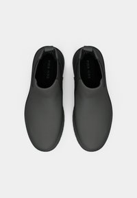 Zapatos negros sin cordones, hechos de un material suave, con punta redondeada y paneles laterales elásticos para facilitar su uso. Suela plana con un diseño minimalista.