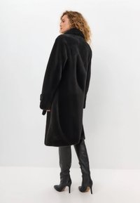 Schwarzer Kunstfellmantel mit hohem Kragen, langen Ärmeln und geknöpften Bündchen, mit einem lockeren Schnitt und weicher Textur. Getragen mit kniehohen Lederstiefeln.