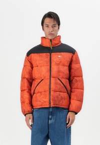 Veste matelassée orange vif avec texture florale, panneau d'épaule noir et fermeture éclair à l'avant. Comprend des poches latérales et un col montant.