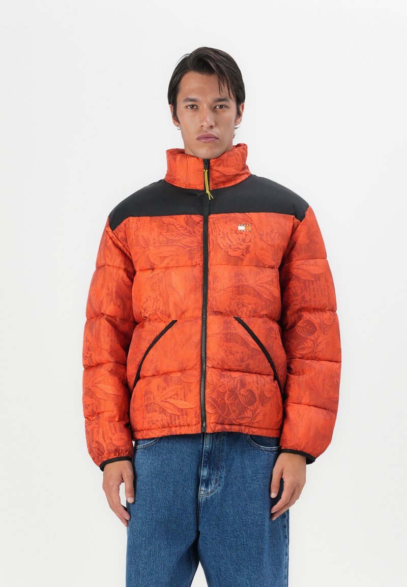 Veste matelassée orange vif avec texture florale, panneau d'épaule noir et fermeture éclair à l'avant. Comprend des poches latérales et un col montant.