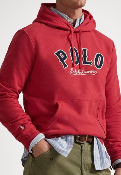 Muškarac nosi crvenu Hoodie Polo Ralph Lauren preko plave prugaste košulje s rukom u džepu zelenih hlača i smeđim remenom.