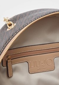 Intérieur d'un sac à main à motif marron avec une fermeture éclair dorée, une doublure intérieure beige, une poche zippée et une étiquette en cuir estampillée "LIU·JO MILANO".