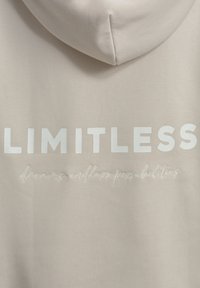 Beige Kapuzenpullover mit weißem Text "LIMITLESS" und kleinerer Schrift darunter, die "Träume, endlose Möglichkeiten" auf dem Rücken liest.